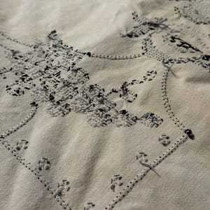 May include: A light beige fabric featuring intricate black and grey embroidery. The needlework displays a detailed floral pattern enclosed within a geometric border, with visible loose threads and textured stitching throughout the design.