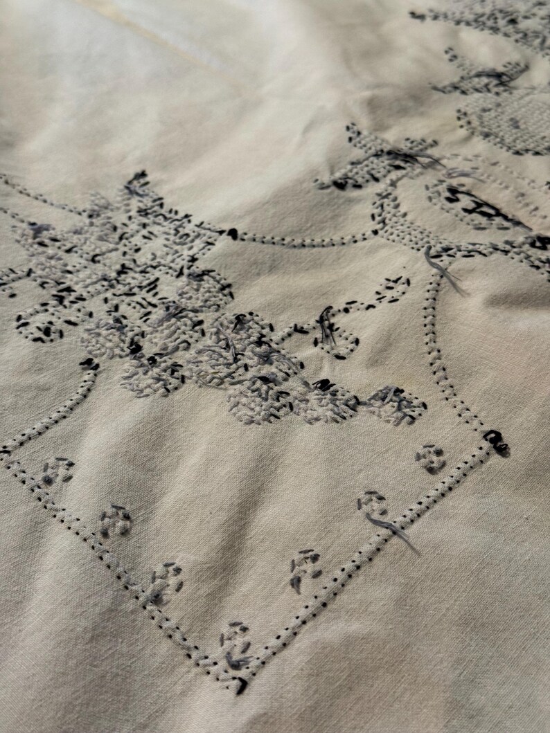 May include: A light beige fabric featuring intricate black and grey embroidery. The needlework displays a detailed floral pattern enclosed within a geometric border, with visible loose threads and textured stitching throughout the design.
