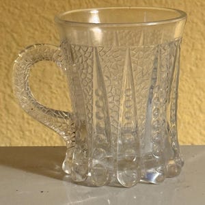 May include: Clear glass mug with a textured, patterned design. The mug features a handle and a decorative base. The glass has a faceted appearance, adding to its vintage charm. A classic piece for serving beverages.