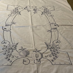 May include: A white rectangular tablecloth featuring a central embroidered border in dark thread. The design consists of a continuous frame decorated with clusters of grapes, various fruits, and leafy vines, creating a symmetrical pattern.