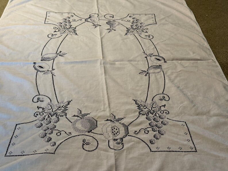 May include: A white rectangular tablecloth featuring a central embroidered border in dark thread. The design consists of a continuous frame decorated with clusters of grapes, various fruits, and leafy vines, creating a symmetrical pattern.