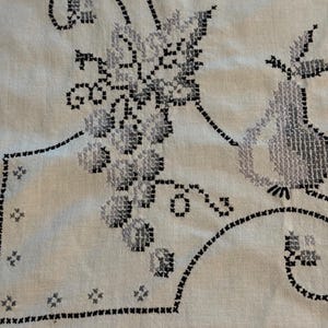 May include: A light-colored fabric featuring intricate cross-stitch embroidery in shades of black and gray. The design displays a cluster of grapes and a pear, accented by decorative scrolling lines and small star-like patterns along the border.