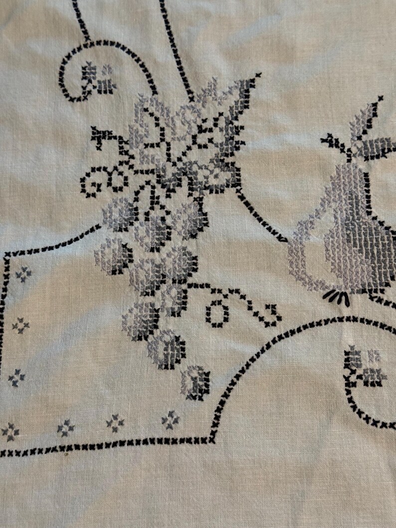 May include: A light-colored fabric featuring intricate cross-stitch embroidery in shades of black and gray. The design displays a cluster of grapes and a pear, accented by decorative scrolling lines and small star-like patterns along the border.