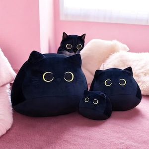 May include: A collection of black cat-shaped plush pillows with yellow eye details. The pillows vary in size, with one cat peeking over the top. The background is a pink surface and a white fluffy pillow.