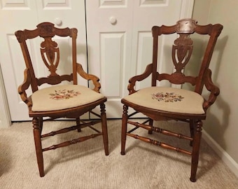 May include: Two antique wooden chairs with ornate backs and armrests. The chairs have beige upholstered seats with floral embroidery. The wood is a warm brown color, and the chairs are set against a white door.