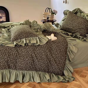 Peut inclure: Un lit avec un ensemble de couette et d'oreillers à motifs floraux. La couette et l'oreiller ont un fond marron foncé avec de petites fleurs blanches. L'ensemble de lit a une bordure à volants vert sauge. Un drap de lit vert sauge est également visible.