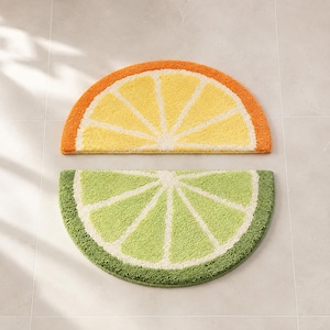 May include: Two semi-circular bath mats shaped like citrus slices. The top mat features a yellow centre with a white segmented pattern and an orange rind border. The bottom mat features a light green centre with a white segmented pattern and a dark green rind border.