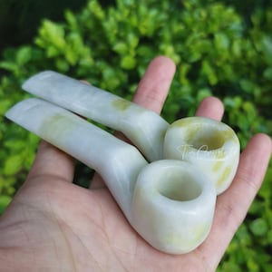 Może przedstawiać: Wyświetlane są dwie biało-żółte fajki z jadeitu. Fajki mają gładką, zakrzywioną konstrukcję, z cybuchem i cybuchą. Fajki są trzymane w dłoni, z zielonym, liściastym tłem.