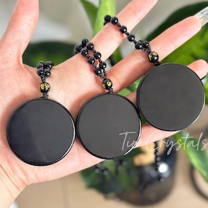 Może przedstawiać: Trzy czarne obsydianowe zawieszki z pozłacanymi napisami na czarnych koralikowych naszyjnikach. Zawieszki są okrągłe i gładkie.