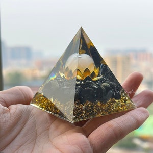 May include: A clear pyramid with gold flakes and black stones inside. A white orb with a gold lotus flower is in the center of the pyramid.