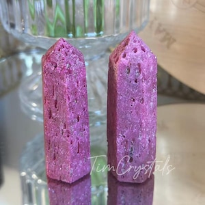 May include: Two vibrant pink crystal towers with textured surfaces. The crystals are rectangular with pointed tops, reflecting on a mirrored surface. The image includes the text "Tim Crystals".
