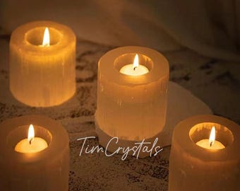 Può includere: Quattro portacandele in selenite illuminati con un bagliore caldo. Ogni portacandele ha un'indentazione circolare per contenere una piccola candela. Il testo "Tim Crystals" è visibile.