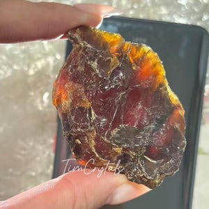 May include: A raw, amber-hued crystal with a dark red core. The crystal is held up, revealing its natural, uneven form and translucent appearance. The text "Tim Crystals" is visible.