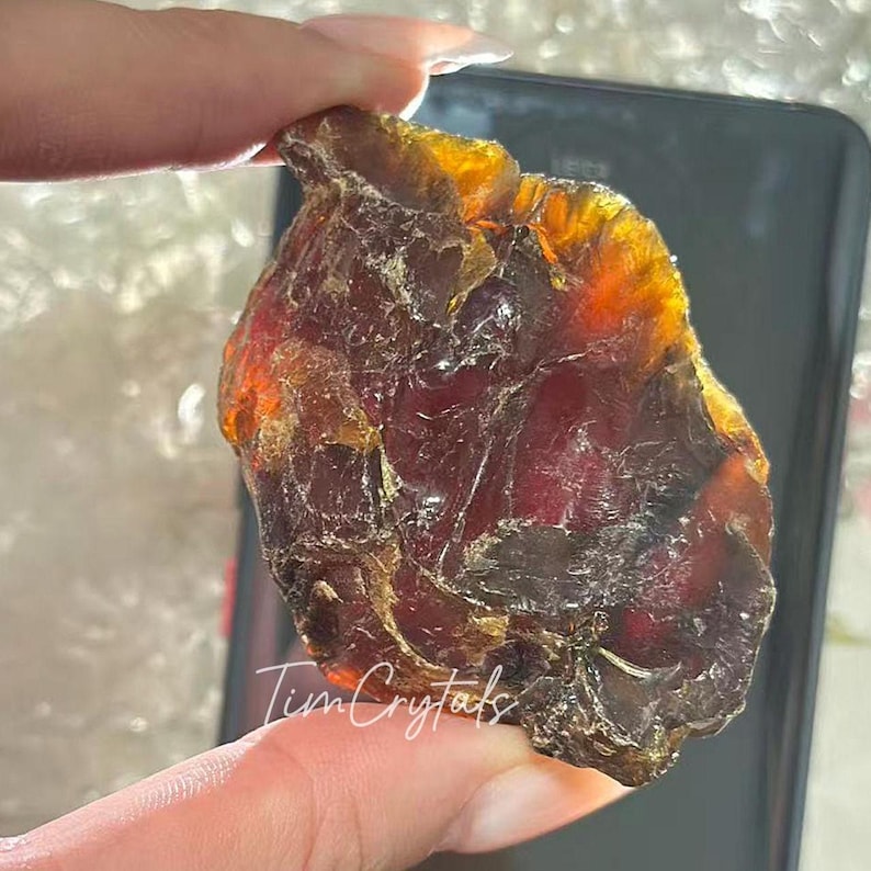 May include: A raw, amber-hued crystal with a dark red core. The crystal is held up, revealing its natural, uneven form and translucent appearance. The text "Tim Crystals" is visible.