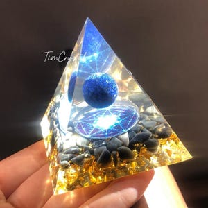 Pode incluir: Uma pirâmide de vidro transparente com uma esfera azul dentro. A pirâmide é preenchida com folha de ouro e pedras pretas e brancas. A esfera é cercada por um círculo prateado com um design geométrico azul e branco.