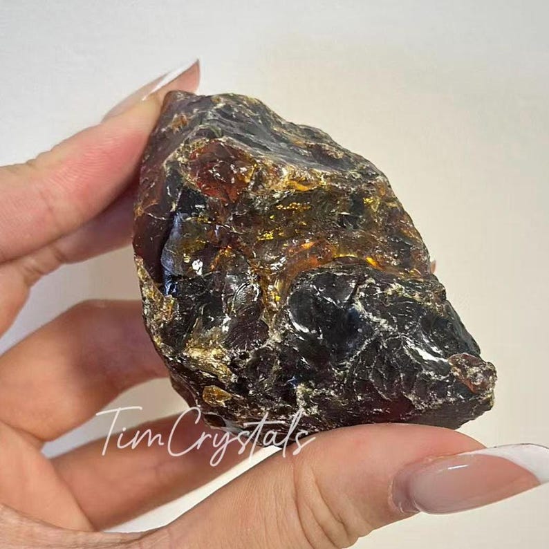 May include: A raw amber crystal, displaying a blend of black and golden-brown colours. The natural stone is held in a hand, revealing its uneven form and textured surface. The image includes the text "Tum Crystals".