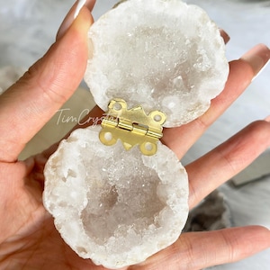 May include: Open, white geode box with a gold-colored hinge. The geode has a crystalline interior and a rough, natural exterior. The box is held open, revealing the interior. The geode is round and has a natural, organic shape.