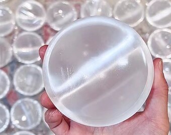 Può includere: Una sfera di selenite bianca con una superficie liscia e lucida. La sfera è tenuta in una mano, e sullo sfondo ci sono altre sfere di selenite.