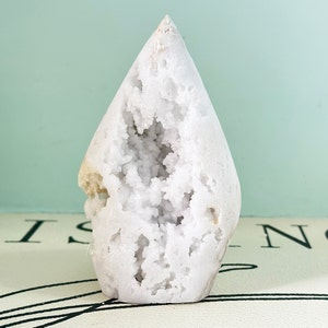 May include: White crystal geode with a large opening revealing a cluster of smaller crystals inside. The geode is shaped like a teardrop and has a smooth, polished surface.