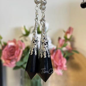 May include: A pair of dangling earrings featuring faceted black stone pendants shaped like elongated points. Each pendant is capped with an intricate silver-toned metal filigree cone and suspended from a silver-toned chain with small beads.