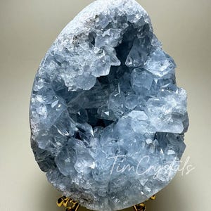 Può includere: Un grande ammasso di cristalli di celestite blu chiaro con un centro vuoto, esposto su un supporto in metallo dorato. Il cristallo ha una texture naturale, ruvida e una varietà di forme e dimensioni.