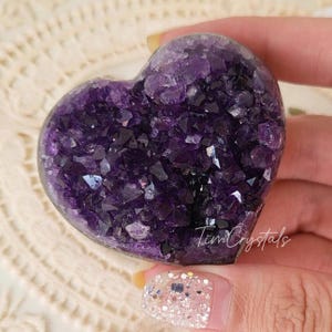May include: A heart-shaped amethyst crystal cluster with a deep purple color. The crystal is polished and has a smooth surface. The crystal is surrounded by a white lace background.