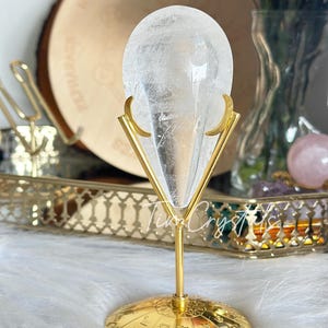 Puede incluir: Una punta de cristal de cuarzo transparente sobre un soporte de metal dorado con una base circular. El soporte tiene un diseño decorativo con forma de media luna a cada lado. El cristal está sobre una superficie blanca y esponjosa.