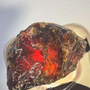 May include: A rough amber gemstone, displaying a rich red and golden colour. The natural stone has a textured surface and internal light refraction. The image includes the text "TimCrystals".