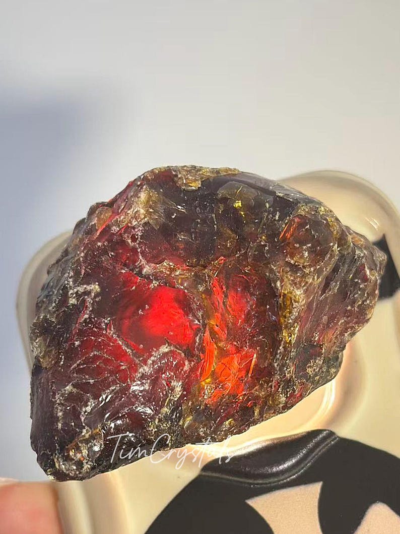 May include: A rough amber gemstone, displaying a rich red and golden colour. The natural stone has a textured surface and internal light refraction. The image includes the text "TimCrystals".