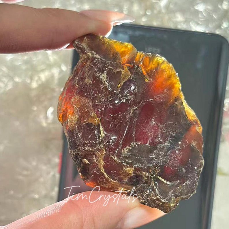 May include: A raw, amber-hued crystal with a dark red core. The crystal has a textured surface and is held in a person's hand. The text "Tim Crystals" is written on the crystal.