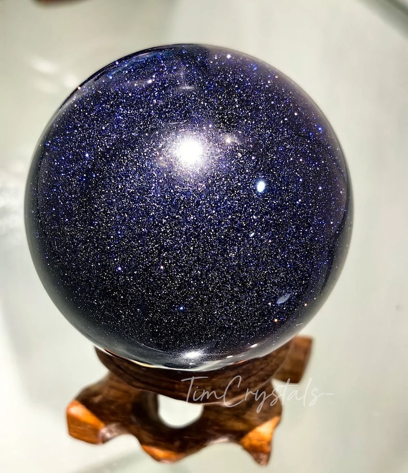 May include: A dark blue, sparkly, polished sphere of blue sandstone on a wooden stand. The sphere is smooth and reflective, with a slight sheen.