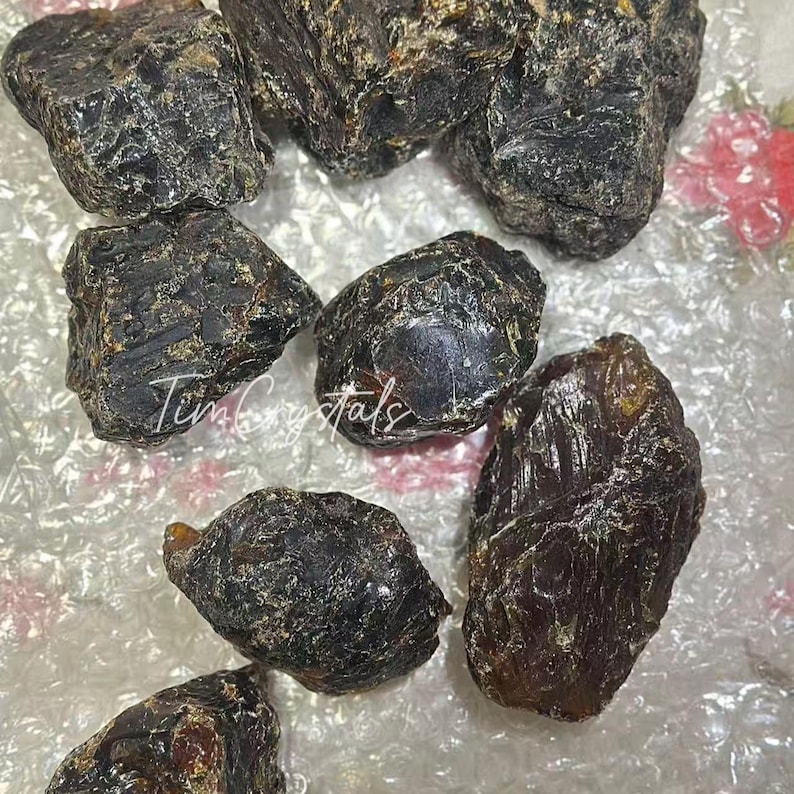 May include: A selection of raw, dark brown amber stones. The amber pieces are of various sizes and shapes, with a glossy, textured surface. The text "Tim Crystals" is visible on the surface.