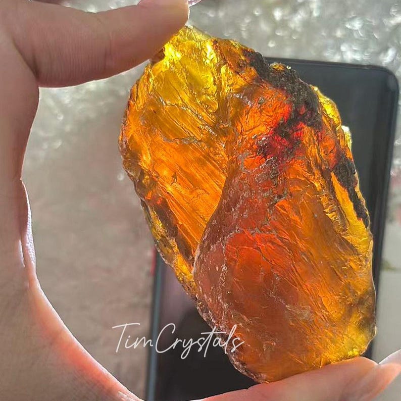 May include: A large, translucent amber-coloured crystal with a rough, textured surface. The crystal is held up, showcasing its golden hues and internal patterns. The text "Tim Crystals" is visible at the bottom.
