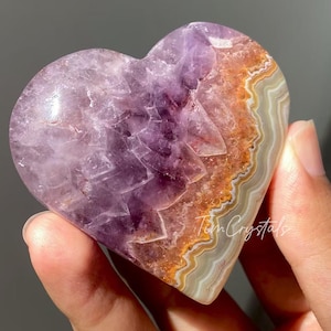 以下が含まれることがあります： 研磨された表面を持つ、ハート型の紫水晶の結晶。 結晶は、紫、オレンジ、白の色合いのグラデーションを特徴としています。 結晶は手で持たれています。