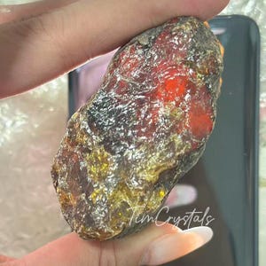 May include: A natural amber gemstone with a rugged, uneven texture. The amber shows a blend of colours, including rich reds, browns, and golden yellows. The image includes the text "Tim Crystals".
