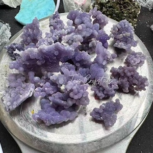 May include: A collection of grape agate clusters with a distinct botryoidal texture and a light purple hue. The mineral specimens are arranged on a circular metal scale, featuring the script text Tim Crystals overlaid across the center of the display.