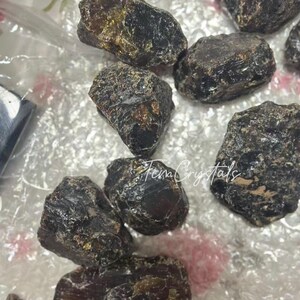 May include: Several dark brown, rough crystals with a glossy surface. The crystals are of various sizes and shapes. The image includes the text "Fena Crystals". These crystals are likely used for metaphysical or decorative purposes.