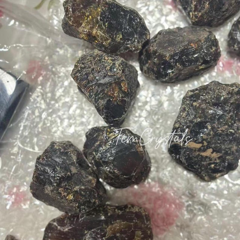 May include: Several dark brown, rough crystals with a glossy surface. The crystals are of various sizes and shapes. The image includes the text "Fena Crystals". These crystals are likely used for metaphysical or decorative purposes.