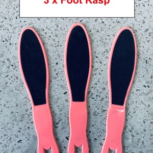 Puede incluir: Tres raspas para pies de color rosa con superficies rugosas negras. Las raspas tienen forma ovalada con un agujero en la parte inferior de cada mango. El texto "3 x Foot Rasp" está escrito en rojo sobre un fondo blanco.