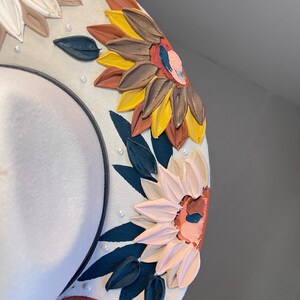 K&ouml;nnte beinhalten: Ein wei&szlig;er Hut mit breiter Krempe, verziert mit gemalten Sonnenblumen und Bl&auml;ttern. Die Sonnenblumen sind in Gelb-, Braun- und Oranget&ouml;nen gemalt. Die Bl&auml;tter sind in Gr&uuml;n- und Schwarzt&ouml;nen gemalt.