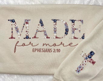 Könnte beinhalten: Beiges Sweatshirt mit floral gesticktem Text "MADE for more" und "Epheser 2,10". Auf dem Ärmel ist ein florales Kreuz gestickt. Die Stickerei zeigt blaue, rote und weiße Blumen.