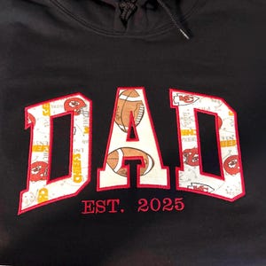 以下が含まれることがあります： 黒いフード付きスウェットシャツに、白の「DAD」が刺繍されています。文字は、フットボールとチームロゴのパターンで埋められています。「EST. 2025」という文字が、文字の下に赤で刺繍されています。