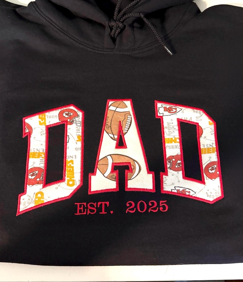 以下が含まれることがあります： 黒いフード付きスウェットシャツに、白の「DAD」が刺繍されています。文字は、フットボールとチームロゴのパターンで埋められています。「EST. 2025」という文字が、文字の下に赤で刺繍されています。