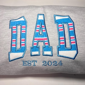 以下が含まれることがあります： グレーのトレーナーに、青とピンクのストライプの刺繍で「DAD」と「EST 2024」の文字が書かれています。