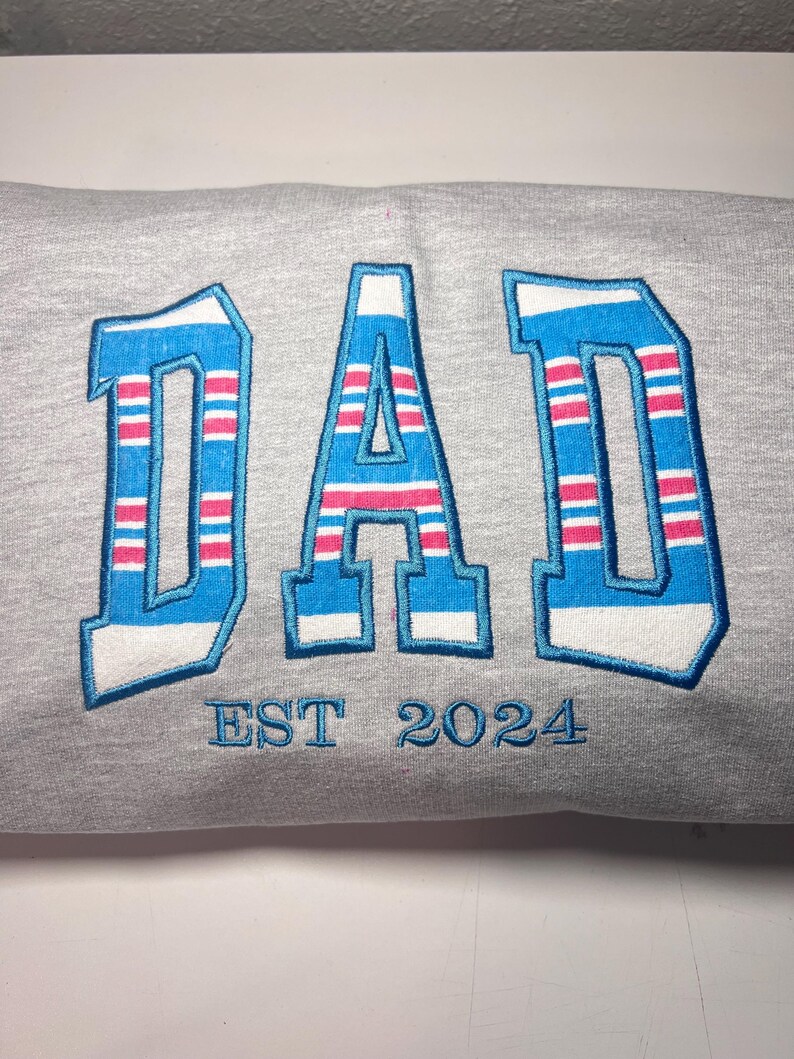 以下が含まれることがあります： グレーのトレーナーに、青とピンクのストライプの刺繍で「DAD」と「EST 2024」の文字が書かれています。