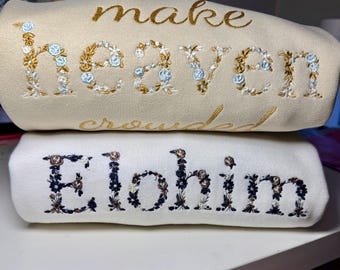 May include: Two stacked sweatshirts. The top one is beige with gold embroidery that reads "make heaven crowded" with floral accents in blue and gold. The bottom sweatshirt is white with "Elohim" embroidered in dark blue and brown floral accents.
