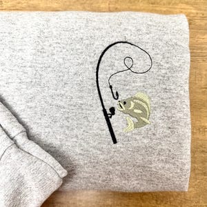 May include: A gray sweatshirt with an embroidered design of a fishing rod and a fish. The fish is biting the hook on the fishing rod.
