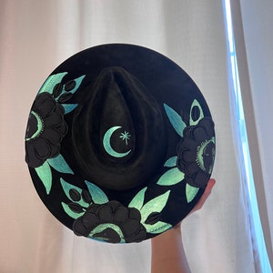 May include: A black felt hat with a crescent moon and star design in teal green. The hat is decorated with black flowers and teal green leaves.
