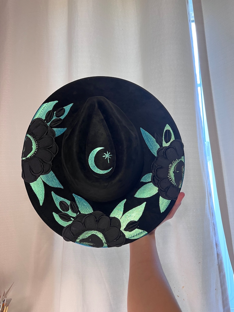 May include: A black felt hat with a crescent moon and star design in teal green. The hat is decorated with black flowers and teal green leaves.