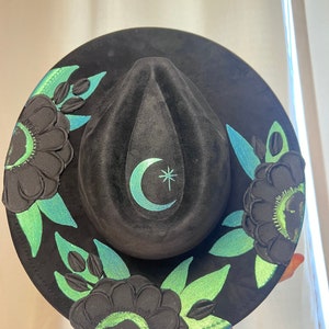 May include: Black wide-brimmed hat with a green and blue floral design. The hat has a crescent moon and star design on the crown.